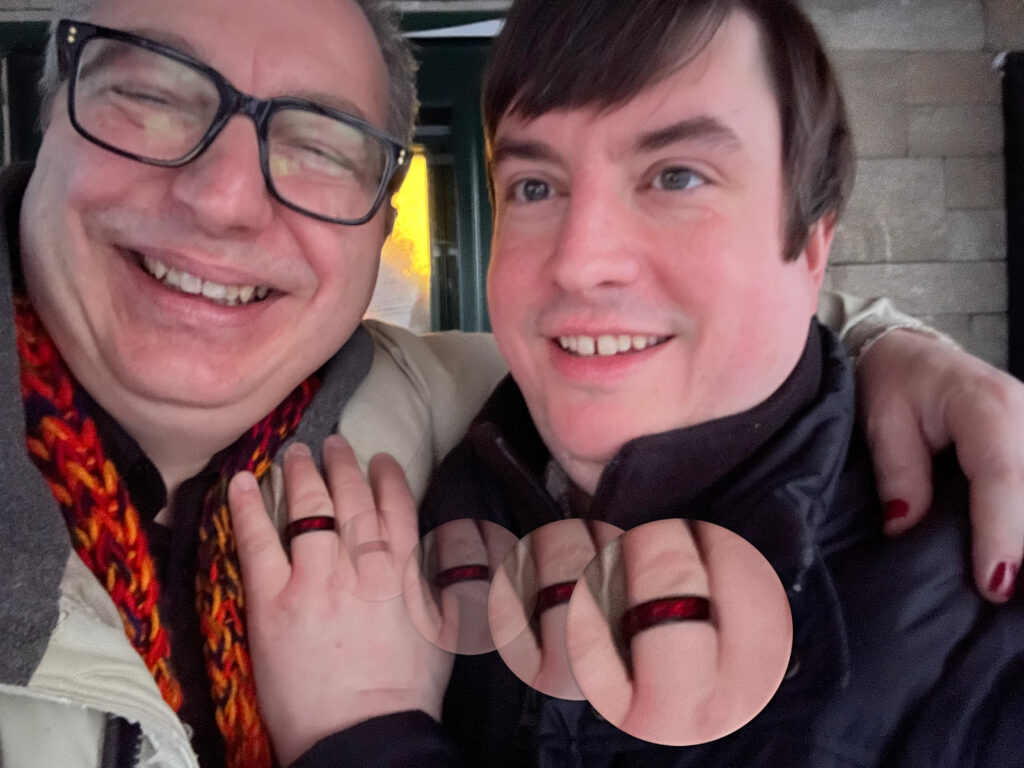 man in glasses with arm around man displaying hand with engagement ring